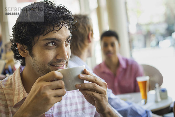 Urbaner Lebensstil. Drei junge Männer um einen Tisch in einem Cafe. Ein Mann nimmt einen Drink aus einer Tasse Kaffee.