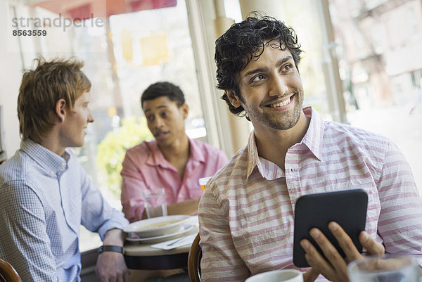 Urbaner Lebensstil. Drei junge Männer um einen Tisch in einem Cafe. Einer hält ein digitales Tablett in der Hand.