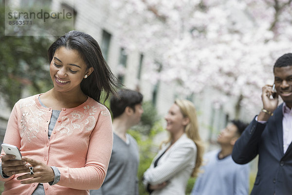 Stadtleben im Frühling. Jugendliche im Freien in einem Stadtpark. Eine Frau in einem rosa Hemd  die ihr Handy überprüft. Vier Menschen im Hintergrund.