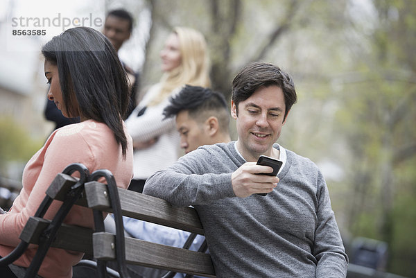 Stadtleben im Frühling. Jugendliche im Freien in einem Stadtpark. Sitzen auf einer Parkbank. Fünf Personen  Männer und Frauen  überprüfen ihre Telefone.
