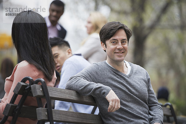 Stadtleben im Frühling. Jugendliche im Freien in einem Stadtpark. Sitzen auf einer Parkbank. Ein Mann lächelt in die Kamera. Vier Menschen im Hintergrund.