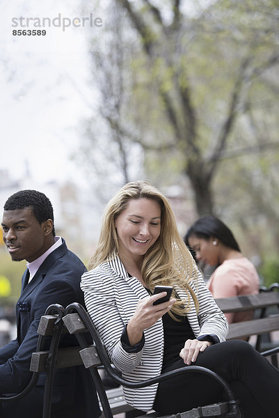 Stadtleben im Frühling. Jugendliche im Freien in einem Stadtpark. Drei Menschen sitzen auf einer Bank. Zwei überprüfen ihre Telefone.