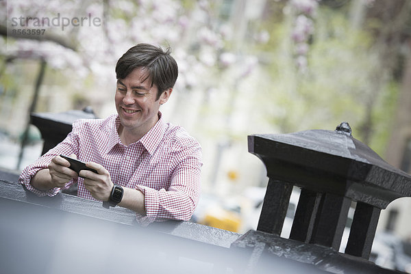 Stadtleben im Frühling. Ein Mann sitzt draußen in einem Stadtpark. Er schaut auf sein Smartphone und lächelt.