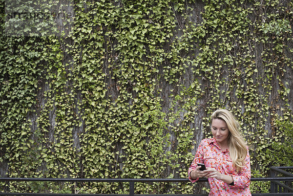 Stadtleben im Frühling. Stadtpark mit einer mit Kletterpflanzen und Efeu bewachsenen Wand. Eine junge blonde Frau mit blonden Haaren  die ihr Smartphone kontrolliert.