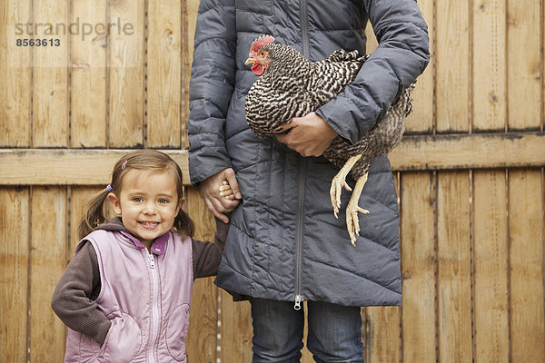 Eine Frau in grauem Mantel  die ein schwarz-weißes Huhn mit rotem Hahnenkamm unter einem Arm hält. Neben ihr ein junges Mädchen  das die andere Hand hält.