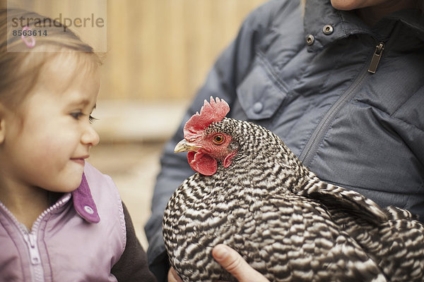 Eine Frau in grauem Mantel  die ein schwarz-weißes Huhn mit rotem Hahnenkamm unter einem Arm hält. Ein junges Mädchen neben ihr  das das Huhn eng an sich hält.