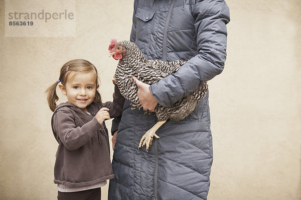 Eine Frau in grauem Mantel  die ein schwarz-weißes Huhn mit rotem Hahnenkamm unter einem Arm hält. Neben ihr ein junges Mädchen  das die andere Hand hält.