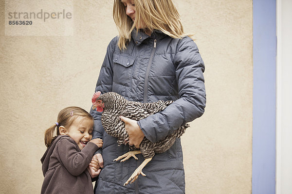 Eine Frau in grauem Mantel  die ein schwarz-weißes Huhn mit rotem Hahnenkamm unter einem Arm hält. Ein junges Mädchen neben ihr  das die andere Hand hält.