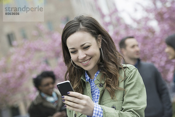 Eine junge Frau sucht im Park auf ihrem Smartphone nach Nachrichten. Im Hintergrund eine Gruppe von Freunden.