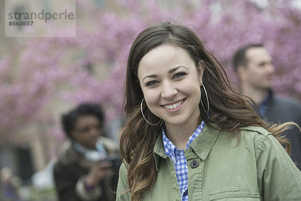 Eine Gruppe von Menschen unter den Kirschblütenbäumen im Park. Eine junge Frau in einem Hemd mit offenem Hals  die lächelt und in die Kamera schaut.
