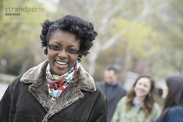 Eine Gruppe von Menschen in einem Stadtpark. Eine junge Frau in einem Mantel mit großem Kragen  die lächelt und in die Kamera schaut.
