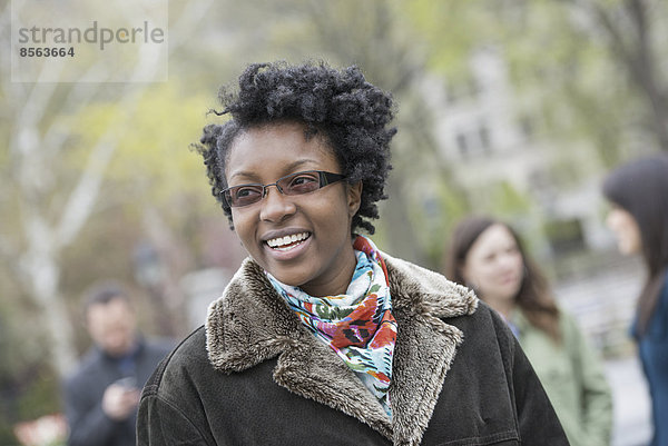 Eine Gruppe von Menschen in einem Stadtpark. Eine junge Frau in einem Mantel mit großem Kragen  die lächelt und in die Kamera schaut.