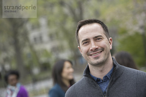 Eine Gruppe von Menschen in einem Stadtpark. Ein Mann in einem grauen Pullover  lächelnd.