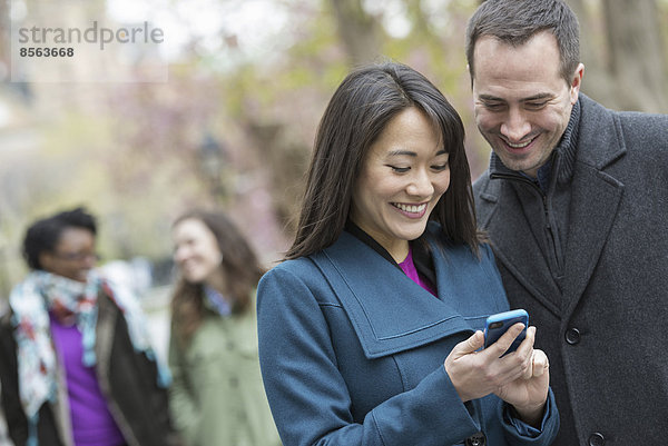 Eine Gruppe von Menschen in einem Stadtpark. Ein Mann in einem grauen Mantel und eine Frau in einem türkisfarbenen Mantel  beide mit Blick auf ein Smartphone.