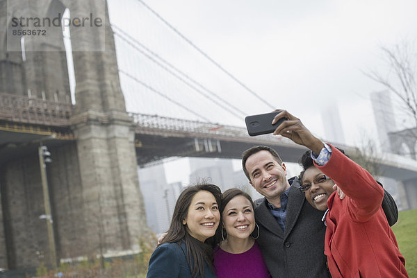 New York Stadt. Die Brooklyn Bridge über den East River. Vier Freunde  die mit einem Telefon ein Foto von sich selbst machen.