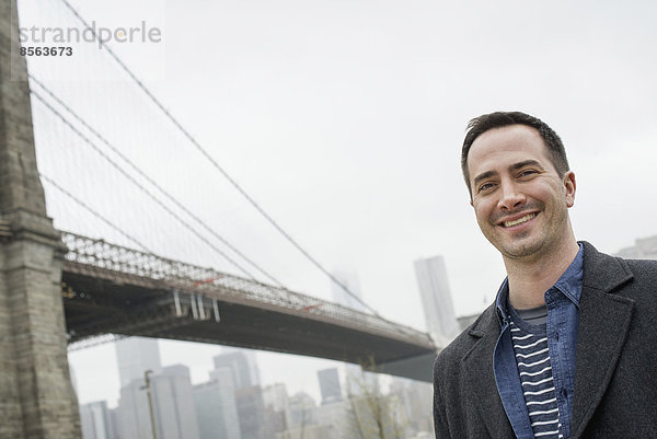 New York City  die Brooklyn Bridge  die über den East River führt. Ein Mann in einem grauen Mantel lächelt in die Kamera.