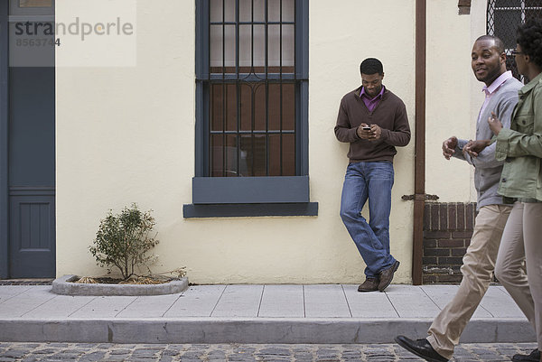 Draußen in der Stadt im Frühling. Ein Paar  das auf dem Bürgersteig geht  und ein Mann  der an einer Wand lehnt und sein Telefon überprüft.