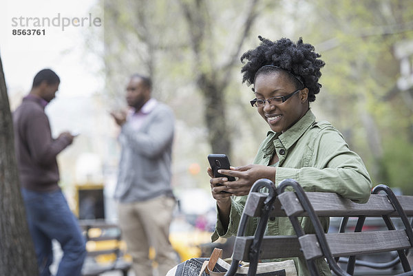 Draußen in der Stadt im Frühling. Ein urbaner Lebensstil. Eine Frau auf einer Bank  die ihr Telefon kontrolliert. Zwei Männer im Hintergrund.