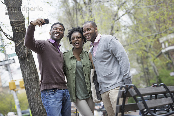 Draußen in der Stadt im Frühling. Ein urbaner Lebensstil. Drei Menschen posieren zusammen und einer macht ein Foto von ihnen mit einem Smartphone.