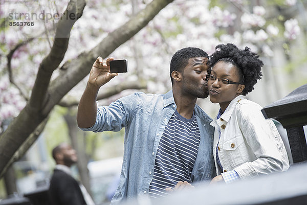Draußen in der Stadt im Frühling. Ein urbaner Lebensstil. Ein Mann küsst eine Frau und macht ein Foto mit einem tragbaren Mobiltelefon.