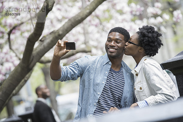 Draußen in der Stadt im Frühling. Ein urbaner Lebensstil. Eine Frau  die einen Mann küsst und mit einem tragbaren Mobiltelefon fotografiert.