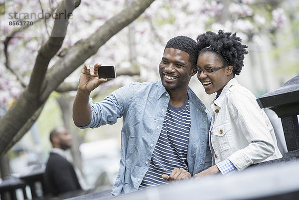Draußen in der Stadt im Frühling. Ein urbaner Lebensstil. Ein Paar nebeneinander  Wange an Wange  das mit einem tragbaren Mobiltelefon fotografiert.