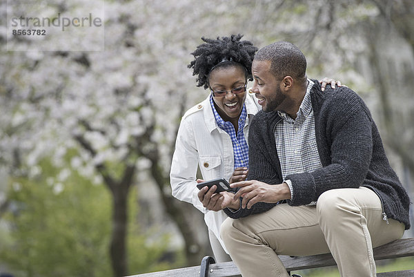 Draußen in der Stadt im Frühling. Ein urbaner Lebensstil. Ein Paar sitzt auf einer Bank  einer hält ein Telefon in der Hand.