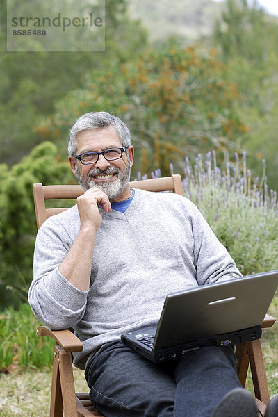Mann sitzt in seinem Garten mit Laptop  lächelt und trägt eine Brille.