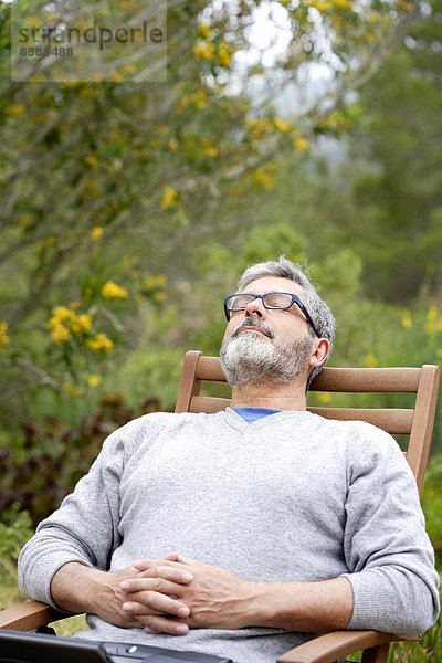 Mann sitzt in seinem Garten mit geschlossenen Augen und lächelndem Gesicht.