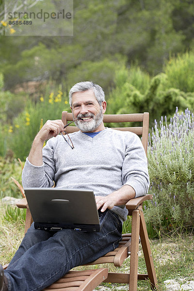Mann sitzt in seinem Garten mit Laptop  lächelnd