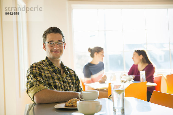 Ein junger Mann sitzt an einer Café-Theke  mit zwei Frauen im Hintergrund.