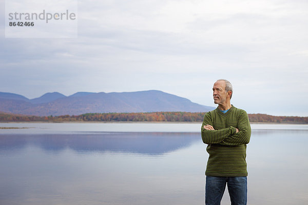 Ein Mann mit verschränkten Armen am Wasser eines flachen  ruhigen Sees in den Bergen.