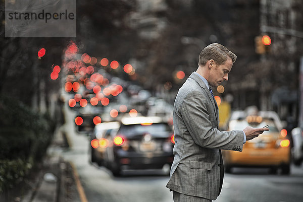 Ein Mann in einem Anzug  der sein Mobiltelefon überprüft  steht in der Abenddämmerung auf einer belebten Straße.