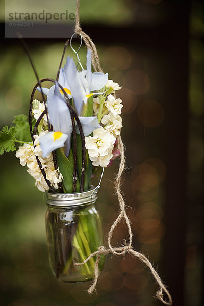 Ein kleines Glasgefäß  das an einem Draht hängt  mit Iris und duftenden Stockblumen. Ein Blumenschmuck.