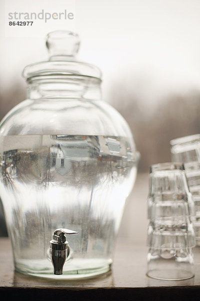 Trinkwasser in einem großen Behälter aus Klarglas. Ein Stapel Gläser. Reines  gekühltes Wasser.