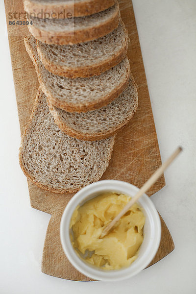 Ein hölzernes Brotbrett  auf dem ein in Scheiben geschnittenes braunes Brot ausgelegt ist. Eine Schale Butter mit einem hölzernen Buttermesser.