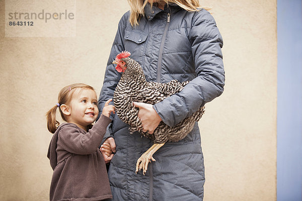 Eine Frau in grauem Mantel  die ein schwarz-weißes Huhn mit rotem Hahnenkamm unter einem Arm hält. Ein junges Mädchen neben ihr  das die andere Hand hält.