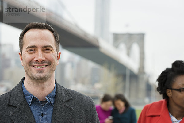 New York City  die Brooklyn Bridge  die über den East River führt. Vier Menschen  zwei Frauen  die auf ein Smartphone schauen  und ein Mann und eine Frau im Vordergrund.