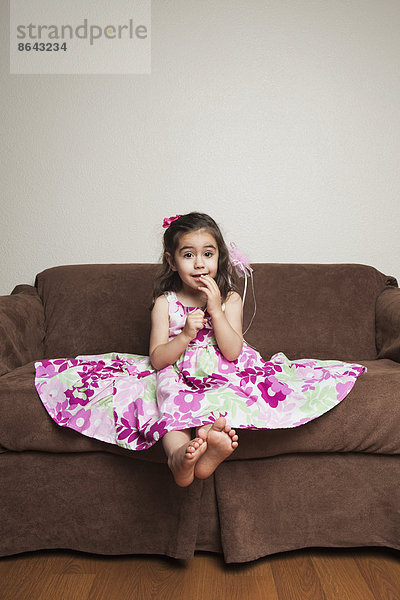 Ein 3-jähriges Mädchen mit langen braunen Haaren in einem rosa geblümten Baumwollkleid mit ausgebreitetem Rock  auf einem braunen Sofa sitzend.