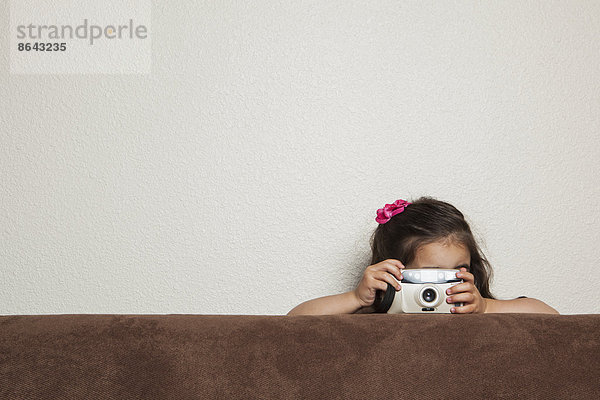 Ein junges dreijähriges Mädchen  das mit einer Spielzeugkamera hinter einem Sofa kauert und ein Foto macht.