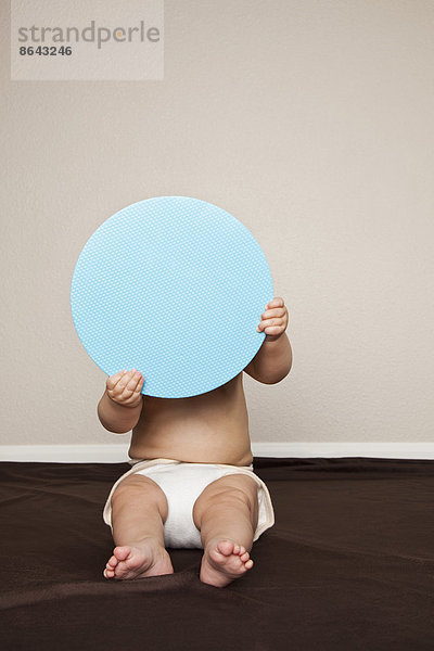 Ein junges 8 Monate altes Baby  das Stoffwindeln trägt und sich hinter einer großen blauen Scheibe versteckt.