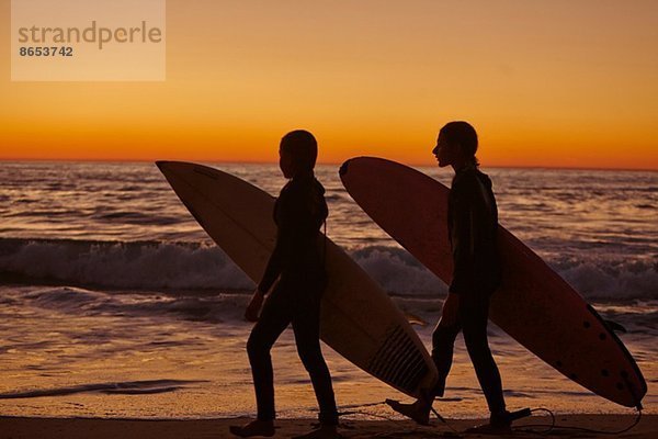 Zwei Mädchen  die bei Sonnenuntergang mit Surfbrettern am Strand spazieren gehen.
