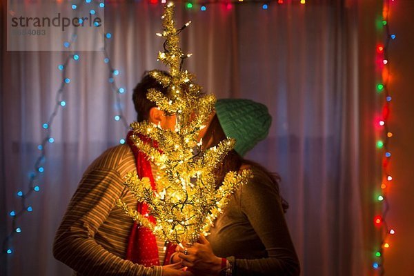 Junges Paar küsst sich hinter dem beleuchteten Weihnachtsbaum