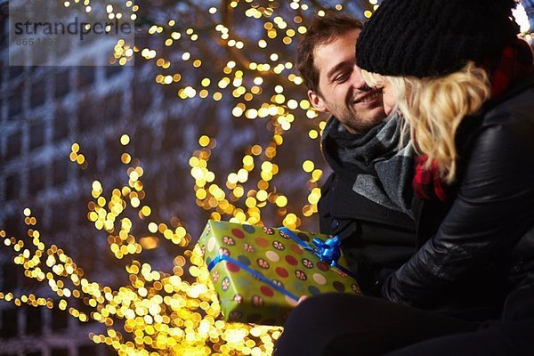 Junges Paar tauscht Geschenke neben den Weihnachtslichtern im Freien aus