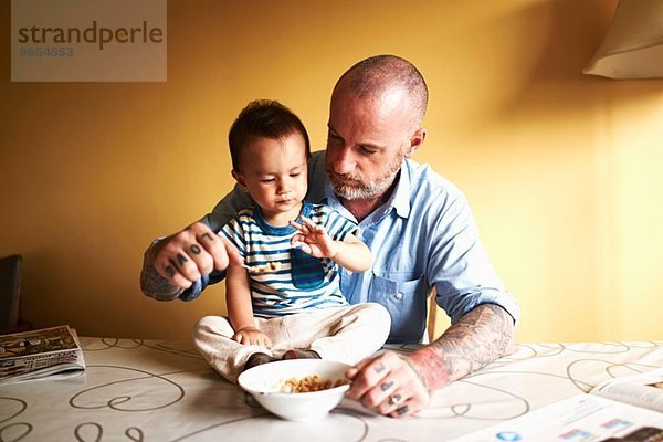 Kleiner Junge sitzt auf dem Tisch und trinkt Müsli mit Vater.