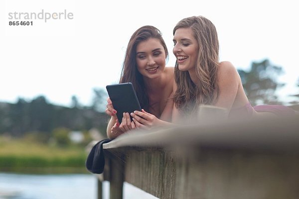 Zwei junge Frauen auf dem Pier am Fluss beim Betrachten des digitalen Tabletts