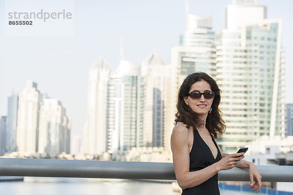 Frau lehnt an Geländer mit Smartphone in der Hand  Wolkenkratzer im Hintergrund