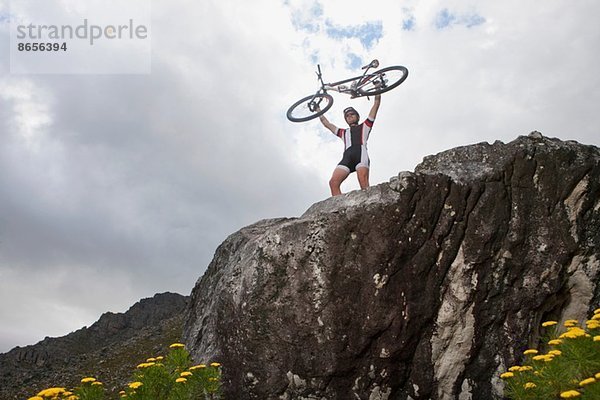 Junger Mann hält Mountainbike auf dem Felsen hoch