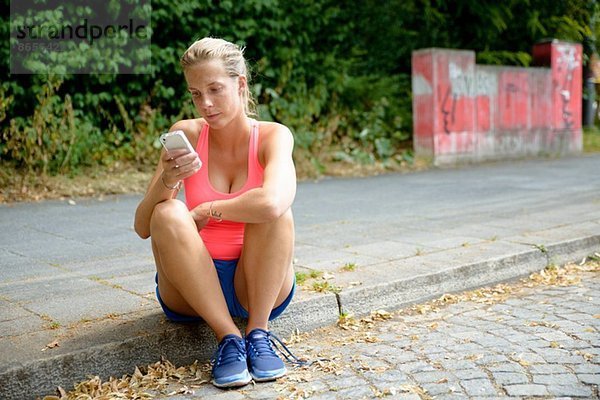 Junge Läuferin auf dem Bürgersteig sitzend mit dem Handy