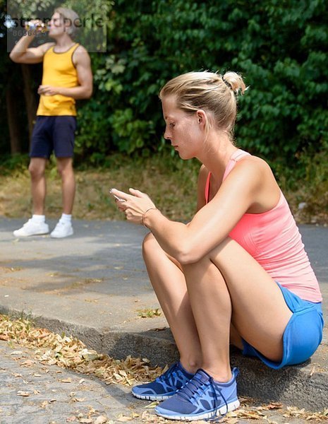 Junge Läuferin sitzt auf dem Bürgersteig und schaut aufs Handy.